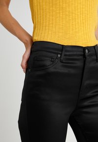 Černé slim-fit džíny s hladkou texturou a jemným leskem. Obsahují standardní přední kapsy a zavírání na horní knoflík, doplněné žlutým žebrovaným topem.