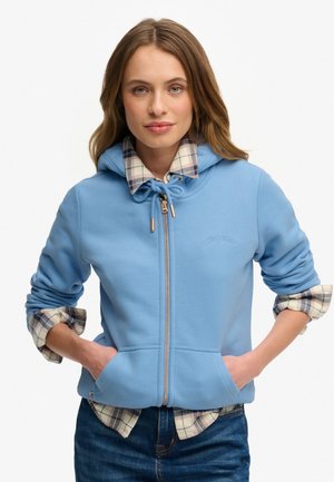 Mujer con cabello castaño claro que lleva una sudadera con capucha con cremallera de color azul claro sobre una camisa de cuadros con las mangas remangadas, manos en los bolsillos de la sudadera.