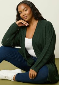 Mörkgrön ribbad cardigan med öppen front, texturerade ärmar och knapppynt. Matchas med en vit topp och mörk denimjeans.