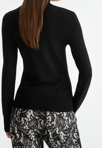 Pull noir à col montant, à manches longues et avec un ourlet côtelé ; porté avec un pantalon noir et blanc à motifs texturés avec un design floral.
