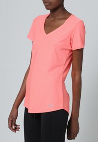 Mujer vistiendo una camiseta de deporte de cuello en V de color coral rosa y leggings negros, de pie contra un fondo gris simple.