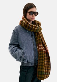 Kvinde iført solbriller med skildpaddeskal, grå denimjakke, sorte bukser og et stort brunt mønstret tørklæde omkring halsen.