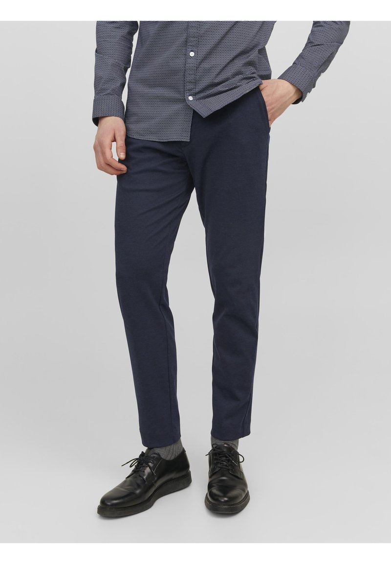 Granatowe spodnie slim-fit wykonane z miękkiego materiału, zestawione z czarnymi sznurowanymi butami i szarymi skarpetkami, na jednolitym jasnym tle.