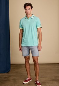 Lichtblauwe polo met contrasterende gele en donkerblauwe kraag en mouwen, gecombineerd met denimshorts en rode sneakers.