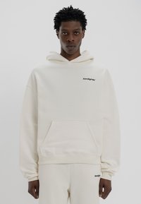 Κρεμ χρώματος φούτερ από μαλακό ύφασμα, με μπροστινή τσέπη και μαύρο λογότυπο στο στήθος. Χαλαρή εφαρμογή με κουκούλα που έχει κορδόνι.