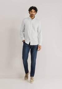Witte button-up shirt met een kraag, gecombineerd met donkerblauwe jeans en beige sneakers, gedragen door een staande mannelijke model. Glad stoftextuur.