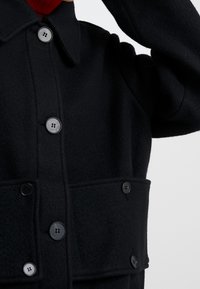 Chaqueta de lana negra con cierre de botones en el frente, cuello puntiagudo y dos bolsillos laterales. La tela tiene una textura suave y un diseño limpio y ajustado.
