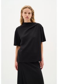 Čierny krátky rukáv top vyrobený z mäkkej bavlnenej látky, so asymetrickým dizajnom a vysokým golierom. Spárovaný s laditou elegantnou sukňou.