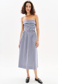 Sininen ja valkoinen ruudullinen olkaimeton mekko, jossa on vyöllinen vyötärö, virtaava midi-pituus ja teksturoitu kangas. Yhdistetty mustiin loafer-kengiin.