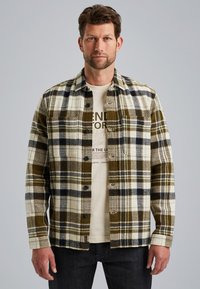 Geruite shirt in gemêleerd groen, beige en marineblauw; gemaakt van gestructureerde stof met een knoopsluiting en twee voorzakken; gedragen over een beige T-shirt.