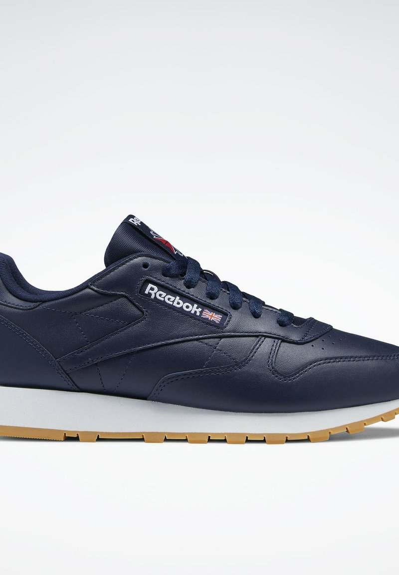 Tamsiai mėlyni odiniai sportbačiai su baltu ir rudu pado, turintys raištelių užsegimą, Reebok ženklinimą ir modernų dizainą.