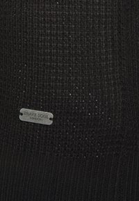 Pull en laine noire avec un motif texturé et une étiquette métallique portant "BRAVE SOUL LONDON" cousue sur le devant.