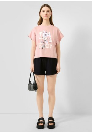 Junge Frau trägt ein pinkes T-Shirt mit Blumenprint, schwarze Shorts, schwarze Sandalen und hält eine kleine schwarze Handtasche vor einem grauen Hintergrund.