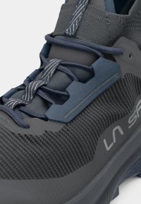 Shoe atlético cinza com uma superfície texturizada, apresentando detalhes em azul, um sistema de atacadores ajustado e um colarinho almofadado para suporte e conforto.