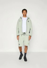 Light green Nike sports jacket with hood, zip pockets, and logo, paired with matching shorts. Black socks and shoes complete the look.
