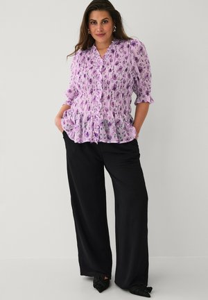 Kobieta w fioletowej kwiecistej bluzce z bufkami na rękawach i czarnych spodniach z szerokimi nogawkami, stojąca z rękami w kieszeniach na jednolitym tle.