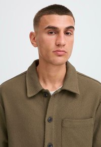 Olive green ribbed jacket with a collared neck, button front, and a single chest pocket; features a textured fabric for added depth.