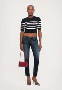Woman standing wearing black and white striped crop top, dark blue jeans, brown heels, holding a small red handbag by her side.