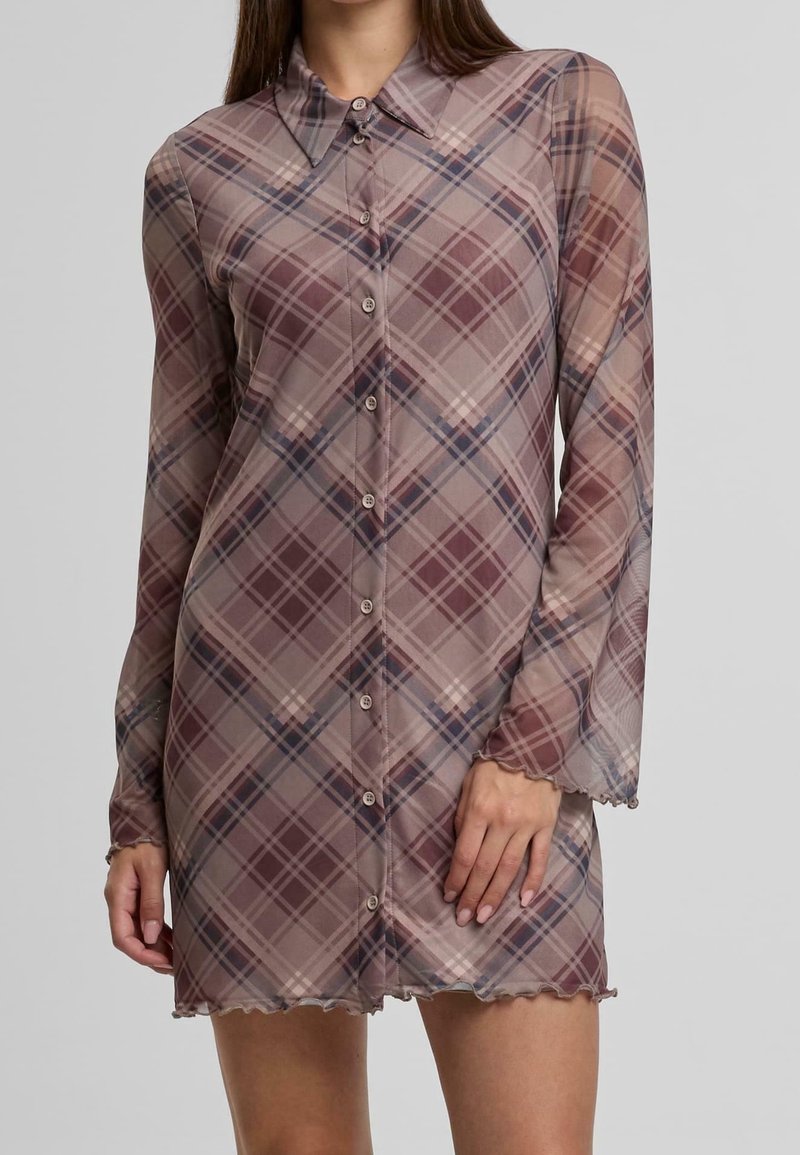 Femme portant une mini-robe à manches longues boutonnée, avec un motif à carreaux marron et violet atténué et un col pointu.