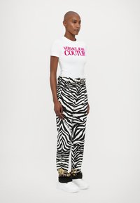 Balts T-krekls ar rozā "Versace Jeans Couture" logo, apvienots ar melnbalti zebras svītrotiem biksēm un baltām masīvām kedām.