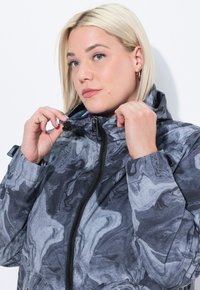 Blond kvinna som justerar huvan på en grå och svart marmorzipjacka mot en vit bakgrund.
