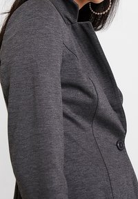 Kvinde iført en tætsiddende mørkegrå blazer med en enkelt sort knap og store hoop øreringe, vist fra siden og op til brystet.