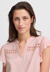 Helle rosa Bluse mit V-Ausschnitt, VolantÄrmeln und besticktem Spitzenbesatz über der Brust, aus weichem, leichtem Stoff gefertigt.