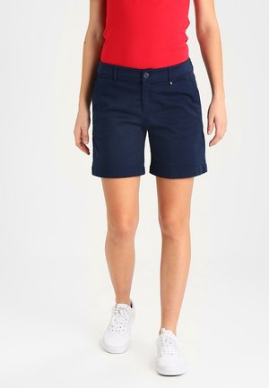 Pantaloni corti in cotone blu navy con chiusura a bottone e orli arrotolati, abbinati a una maglietta rossa e sneakers bianche. Design semplice e casual.
