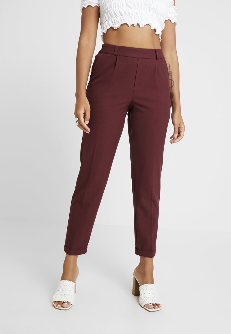 Femme portant un pantalon bordeaux taille haute, fuselé avec des ourlets retournés, des talons blancs tissés à bout ouvert, et un top court blanc avec des bords volantés.