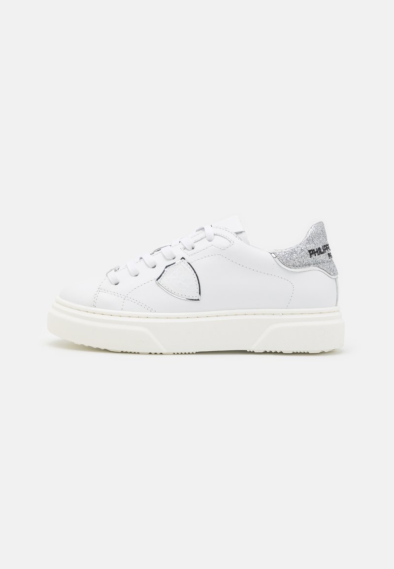 Zapatillas de cuero blancas con punta redonda, que presentan un acento en el talón de color plateado brillante y una suela de goma texturada. Los cordones son blancos.