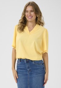 Blouse jaune à manches courtes avec un décolleté en V et des détails froncés, associée à une jupe en denim. Le haut a une texture douce et une coupe détendue.