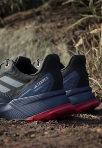 Czarno-niebieskie buty sportowe z teksturowaną cholewką, różowymi akcentami i czerwoną gumową podeszwą. Charakteryzują się wyraźnym logo i warstwowym wzornictwem.