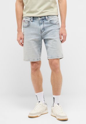 Man wearing light blue denim shorts, white crew socks with black text, beige and white sneakers, and a beige t-shirt, standing against a white background.