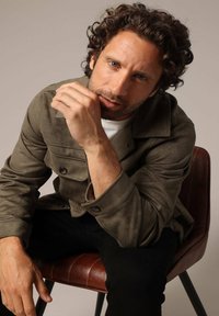 Homme aux cheveux bouclés et à la barbe de trois jours, portant une veste olive et un pantalon noir, assis sur une chaise marron, penché en avant avec une main près du visage.