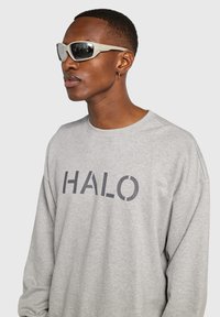 Ljusgrå sweatshirt med "HALO" i mörkgrå bokstäver. Har rund halsringning och långa ärmar. Modellen bär beige solglasögon med reflekterande linser.