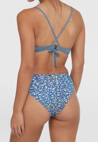 Kék bikini felső, keresztezett pántokkal és megkötős zárással. Az alsó részen kék, sárga és fehér absztrakt formákból álló, színes mintázat található.