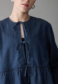 Femme portant un haut en denim bleu foncé avec des nœuds noués au niveau du col et de la taille, accompagnée de boucles d'oreilles en argent, sur fond gris.