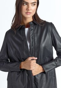 Sort læderjakke med knapper, glat tekstur, klassisk krave og sidesømme, båret over en hvid top.