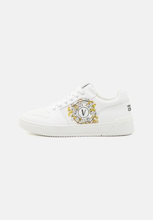 Valkoinen nahkainen tennari, jossa on koristeellinen keltainen kukkakirjailu ja logotieto sivulla. Teksturoitu varvasosa, kumipohja ja nauhavarustus.