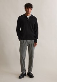 Suéter preto de malha com colarinho, detalhes em botão, combinado com calças plissadas cinza claro e sapatos de couro pretos. Fundo simples.