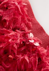 Červená látka s plyšovou textúrou podobnou periu a lesklými flitrovými akcentmi, vytvárajúca vrstvený a textúrovaný vzhľad.