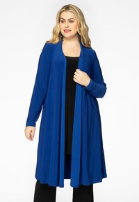 Langer blauer Cardigan aus einem glatten Stoff, mit offener Front und langen Ärmeln. Über einem schwarzen Oberteil getragen, mit einer fließenden Silhouette.
