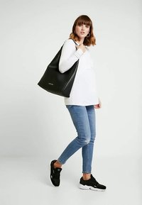 Mulher vestindo uma blusa de manga longa branca, jeans azuis e ténis pretos, carregando uma grande bolsa preta de ombro, sobre um fundo branco liso.