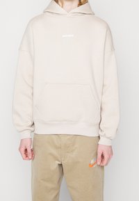 Personne portant un sweat à capuche beige avec une poche avant et un pantalon Nike beige avec un logo swoosh orange sur la cuisse gauche.