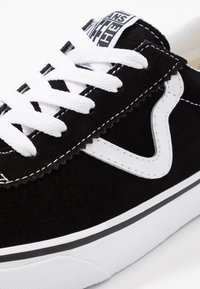 Black suede sneaker with white laces and a contrasting white logo. The sole is flat with a black and white stripe. Background is plain white.