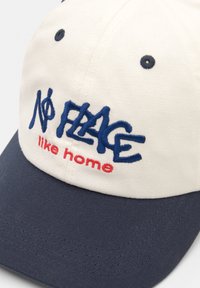 Casquette de baseball en coton avec une couronne crème et un visière bleu marine. Présente un texte brodé "Aucun endroit comme chez soi" en bleu et rouge.