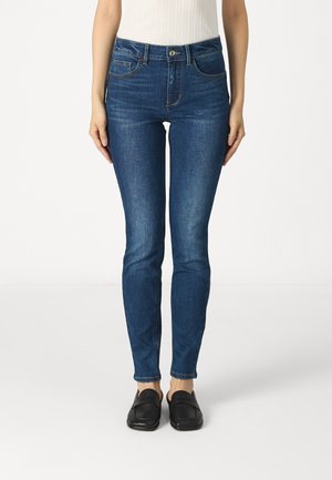 Jeans skinny di colore blu scuro in denim, con vita media, tasche frontali e una vestibilità strutturata fino alla caviglia, abbinati a scarpe nere.