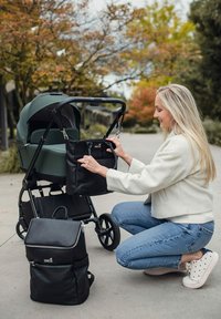 Černá kožená přebalovací taška připnutá na zelený kočárek, na zemi je černý batoh. Scéna se odehrává venku s stromy v pozadí.