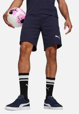 Uomo con pantaloncini navy Puma, calzini neri Puma con strisce bianche, scarpe da ginnastica navy Puma, che tiene un pallone da calcio bianco e rosa.