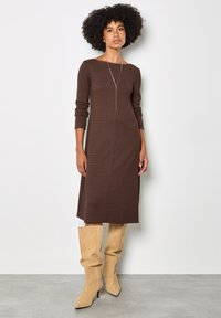Brown long-sleeve midi dress with ribbed texture, round neckline, and side slit, paired with cream knee-high boots on a neutral background.